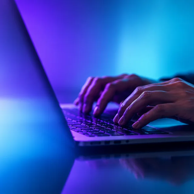 Hands typing on a laptop lit in violet and cyan